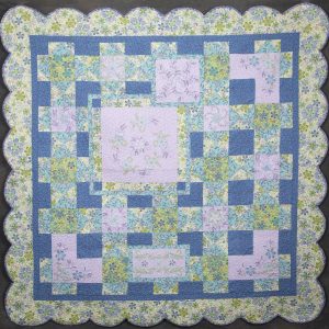 Quilting - Chloe - quilt made of various blue and pink patterned fabrics with a floral design and cut into square shapes and quilted together to create a patterned grid of squares. The quilt is finished with a scalloped edge.