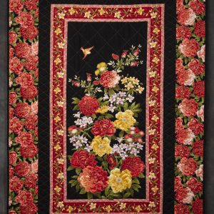 Quilting - Floral - quilt made of a vibrant colored fabric with large red and pink flowers (perhaps roses) printed on it as the border of the quilt and in the center of the quilt is an image of a spray of the same flowers used in the border and accented with sprigs of smaller yellow and pink flowers with a small yellow bird in flight at the top. The center spray of flowers is framed with a black fabric border the connects to the colorful floral border around the quilt.