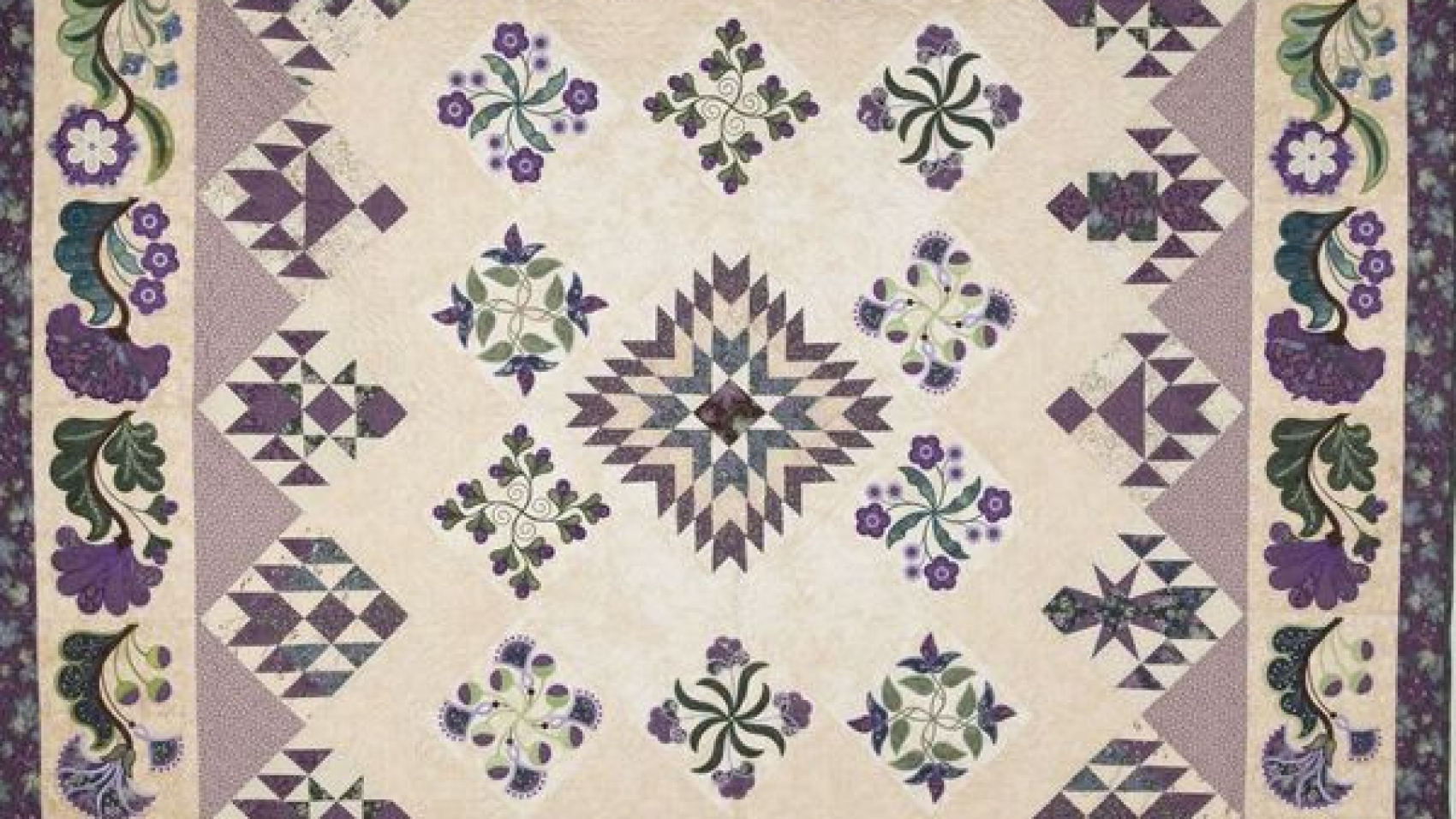Quilt featuring purple flowers and purple and green shapes on an off-wite and lavendar background.