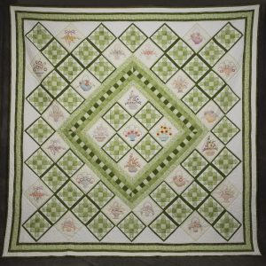 Quilting - Kaufman Panels - quilt in a varitey of green printed fabrics on a white background arranged in different configurations and pieced together. The quilt also features blocks of fabric with baskets of flowers on them and the quilt is framed in a green floral patterned material.