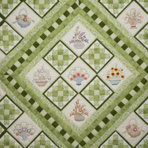 Quilting - Kaufman Panels - Detail - quilt in a varitey of green printed fabrics on a white background arranged in different configurations and pieced together. The quilt also features blocks of fabric with baskets of flowers on them and the quilt is framed in a green floral patterned material.