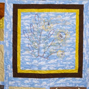 Quilting - Thats Kansas - Detail - quilt of multicolored blocks with a variety of shapes surrounding a larger center block with a blue patterned fabric background with a stitched design in the center depicting sunflowers and cattail plants.