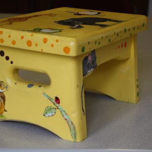 Sculpture 3D - Animal Stool - Detail - small step stool painted a yellow color with a variety of animals painited on it. The top of the stool features a painted green frog with red eyes, a large gray elephant with tusks, a balck and white penguin and a golden giraffe.