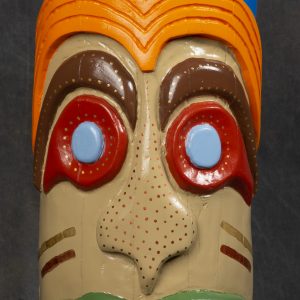 Sculpture 3D - Here Comes the Sun - sculpture carved on a wooden cylinder that resembles a totem pole with brightly colored eyes outlined in red with orange hair and blue top of the pole with yellow sun. The image on the pole is showing its very large teeth outlined with green lips.