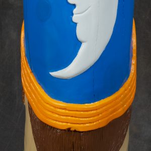 Sculpture 3D - Here Comes the Sun - Detail - sculpture carved on a wooden cylinder that resembles a totem pole with brightly colored eyes outlined in red with orange hair and blue top of the pole with yellow sun. The image on the pole is showing its very large teeth outlined with green lips
